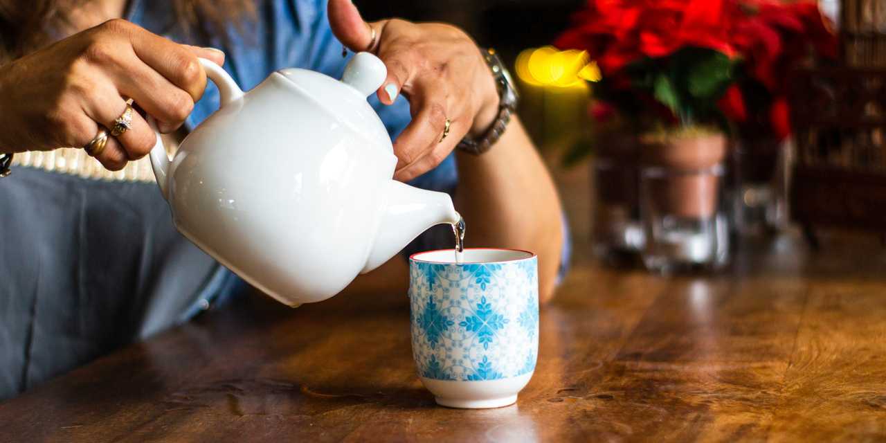 Henkilö kaatamassa juomaa kannusta juomamukiin. Taustalla punaisia kukkia. 