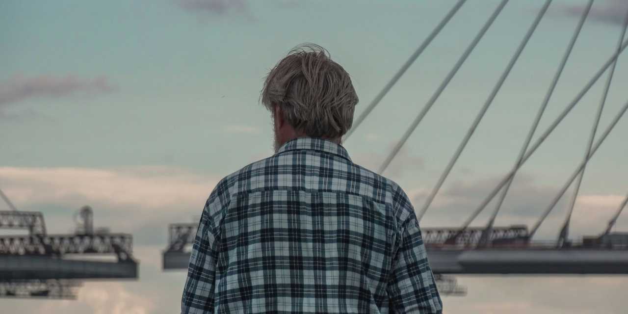 En äldre man med rutig skjorta tittar ut över vattnet med blå himmel och moln.  